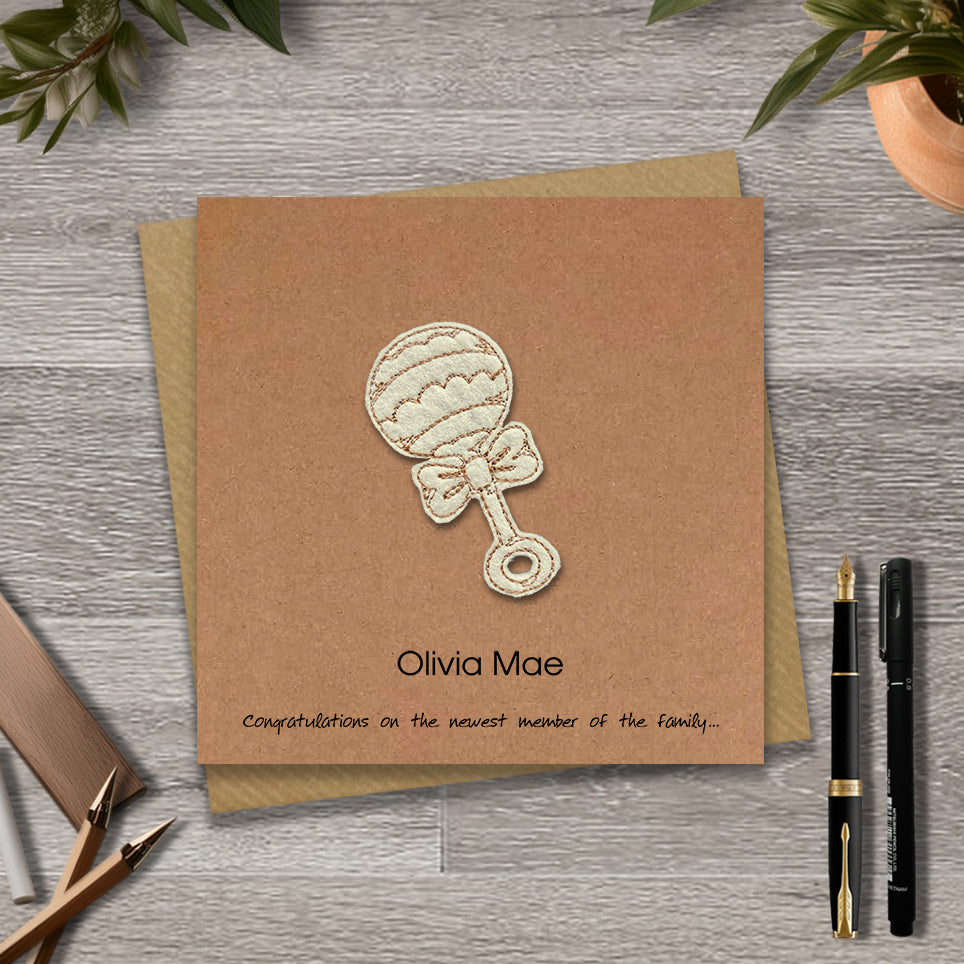 Kraft Baby girl card with embroidered baby's rattle design and personalised text