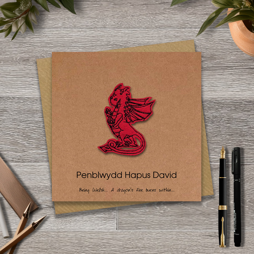 Brown birthday card with red dragon design and 'Penblwydd Hapus David' text on a wooden surface.
